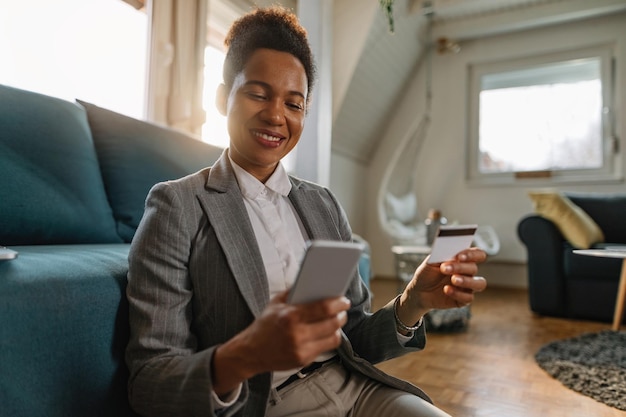 Felice donna d'affari afroamericana che utilizza la carta di credito mentre effettua l'ebanking su smartphone a casa