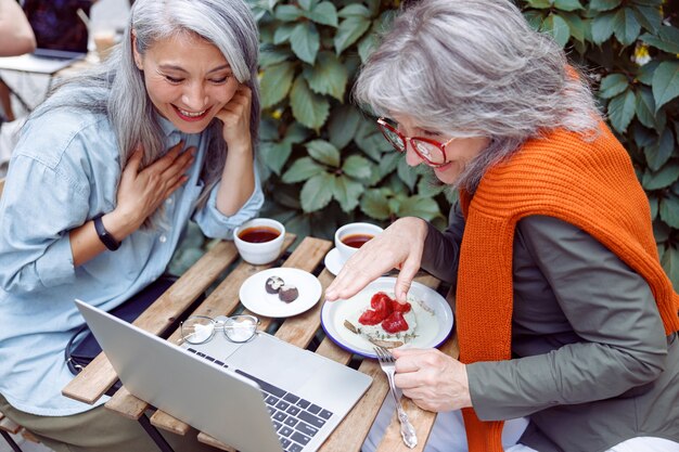 Felice donna dai capelli grigi con un amico guarda video tramite laptop al tavolo sulla terrazza del caffè all'aperto