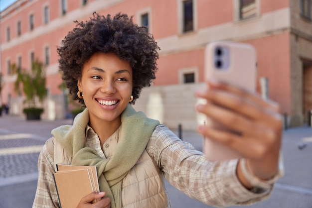 Felice donna dai capelli ricci fa selfie tramite smartphone sorride ampiamente tiene i taccuini indossa camicia e maglione legati sopra il collo sorride ampiamente passeggiate all'aperto ha un'espressione allegra Utente della tecnologia