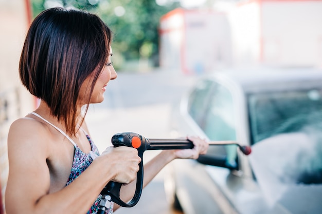 Felice donna di mezza età che lava l'auto alla stazione di autolavaggio utilizzando una macchina per l'acqua ad alta pressione