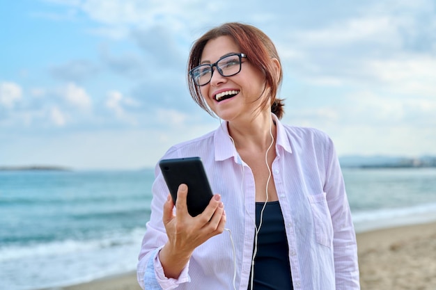 Felice donna di mezza età in vacanza al mare che si gode in cuffia con lo smartphone