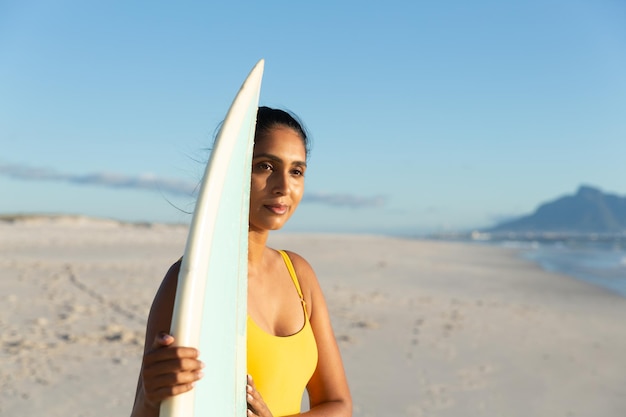 Felice donna di razza mista che porta la tavola da surf sulla spiaggia. sano tempo libero all'aria aperta in riva al mare.