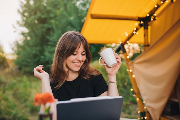 Felice donna freelance che utilizza laptop e bere caffè in un'accogliente tenda glamping in una giornata di sole Tenda da campeggio di lusso per vacanze estive all'aperto e vacanze Concetto di stile di vita