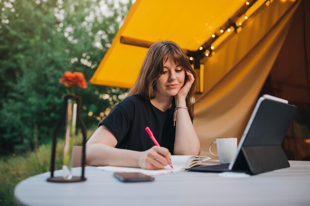 Felice donna freelance che utilizza laptop e prendere appunti seduti su un'accogliente tenda glamping in una giornata di sole Tenda da campeggio di lusso per vacanze estive all'aperto e vacanze Concetto di stile di vita