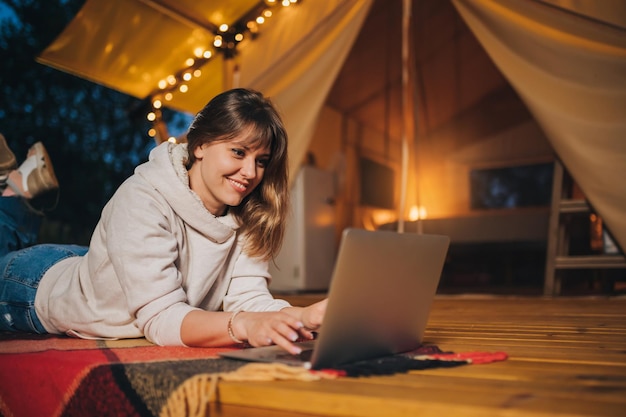 Felice donna freelance che utilizza un laptop su un'accogliente tenda glamping in una sera d'estate Tenda da campeggio di lusso per vacanze all'aperto e vacanze Concetto di stile di vita