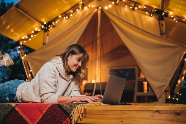 Felice donna freelance che utilizza un laptop su un'accogliente tenda glamping in una sera d'estate Tenda da campeggio di lusso per vacanze all'aperto e vacanze Concetto di stile di vita