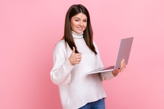 Felice donna freelance lavora sul computer portatile guardando la fotocamera che mostra il pollice in su piace il suo lavoro indossando un maglione bianco in stile casual Studio indoor girato isolato su sfondo rosa