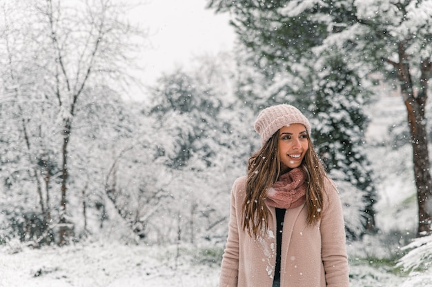 Felice donna in abiti caldi in piedi nei boschi innevati e distogliere lo sguardo mentre si gode il fine settimana invernale