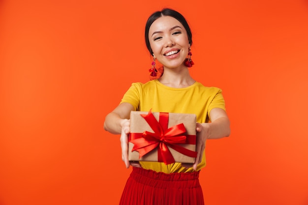 Felice donna ispanica 20s vestita con gonna sorridente e tenendo il regalo di compleanno con fiocco isolato su muro rosso