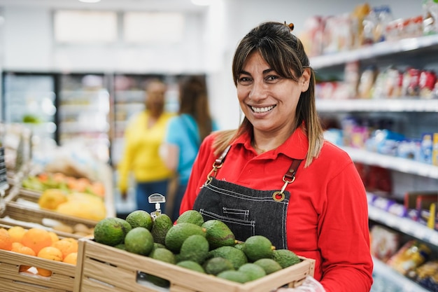 Felice donna latina che lavora all'interno del supermercato mentre tiene una scatola di legno con avocado