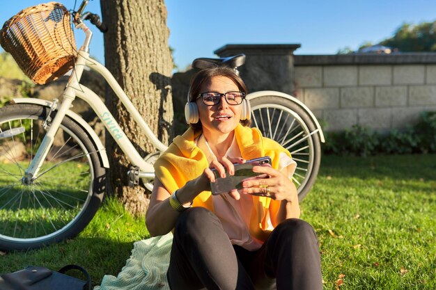 Felice donna matura con le cuffie seduto nel parco cantando e ascoltando musica