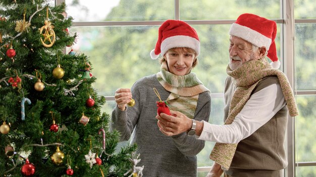 Felice donna matura e uomo anziano in cappelli di Babbo Natale sorridenti e appesi baubles su conifere mentre si preparano per la celebrazione del Natale a casa