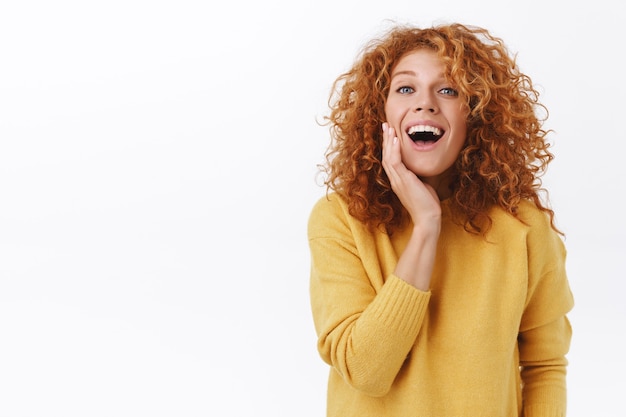 Felice donna rossa sorridente con i capelli ricci, indossa un maglione giallo, ride e tocca il viso sorpreso e contento, felice di vedere che si è sbarazzato dell'acne, soddisfatto dopo la procedura di cosmetici, muro bianco