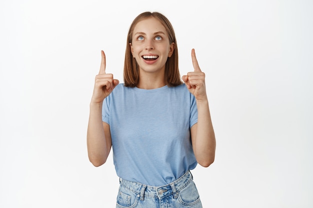 Felice donna sognante con gli occhi azzurri, denti bianchi sorridenti, puntando le dita verso l'alto e guardando affascinata dalla pubblicità, controllando l'offerta promozionale, in piedi con una maglietta blu sul muro bianco.