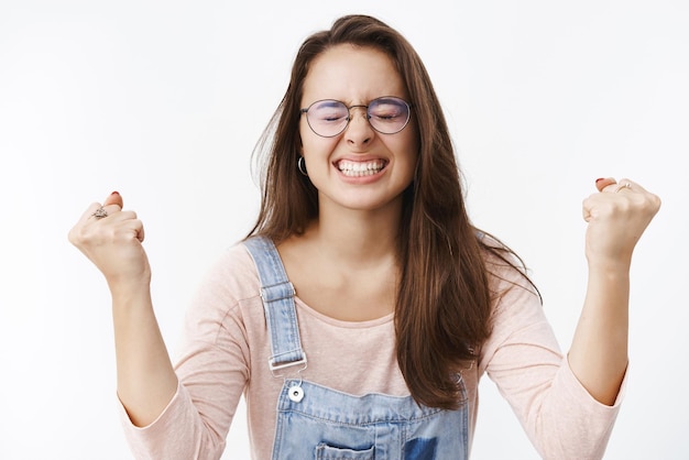 Felice donna sollevata che trionfa mentre finalmente ottiene l'obiettivo o il successo stringendo i denti deliziati alzando i pugni chiusi nella vittoria e nella celebrazione essendo felice di realizzare il sogno su sfondo grigio