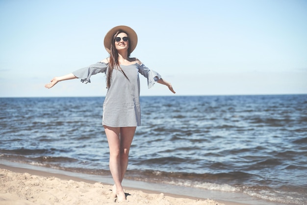 Felice donna sorridente in beatitudine libera sulla spiaggia dell'oceano in piedi con le mani aperte. Modello femminile castana in occhiali da sole e cappello che gode della natura durante le vacanze di viaggio vacanza all'aperto, piano medio.