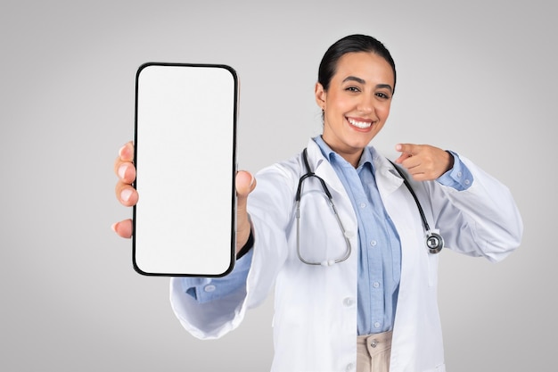 Felice dottoressa latina che mostra il sito web della clinica pubblicitaria sullo schermo del cellulare vuoto o medico