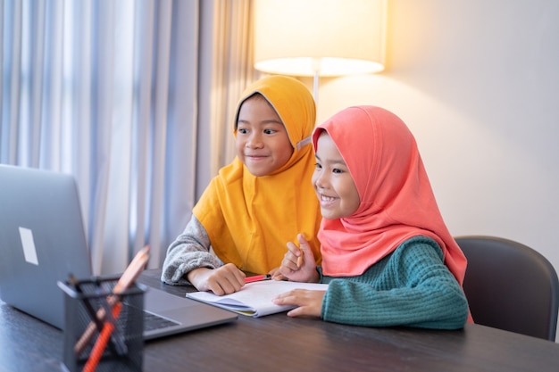 Felice due bambini musulmani che indossano sciarpa per la testa sorridente mentre si utilizza il computer portatile a casa