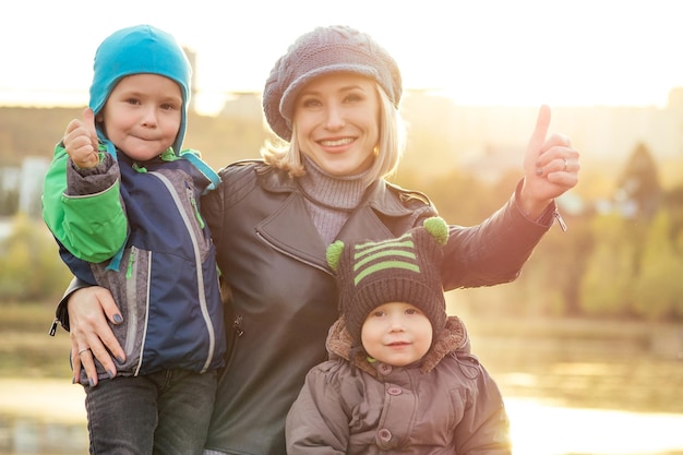 Felice e bella donna bionda con il cappello che gioca due bambini in una giacca calda che lo tiene in braccio nel parco autunnale sullo sfondo del lago madre elegante con un bambino