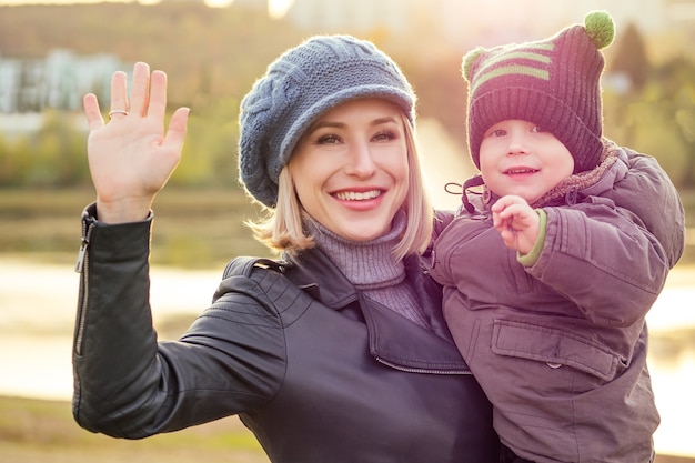 Felice e bella donna bionda in cappello che bacia il figlio del bambino in una giacca calda che lo tiene tra le braccia nel parco autunnale.