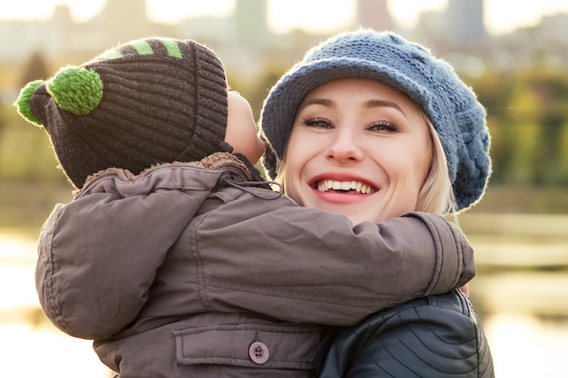 Felice e bella donna bionda in cappello che bacia il figlio del bambino in una giacca calda che lo tiene tra le braccia nel parco autunnale.