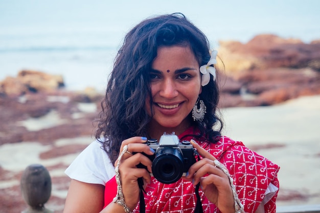 Felice e bella donna indiana nel tradizionale sari rosso alla moda che tiene la fotocamera in mano in posa per il fotografo sulla spiaggia paradiso tropicale mare oceano