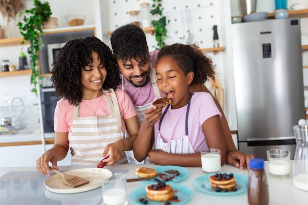 Felice e positivo giovane famiglia sciocco con piccola figlia in cucina che prepara i pancake hanno gustosa colazione posa alla fotocamera bere latte fresco mangiare frittelle