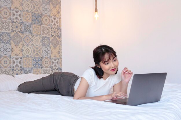 Felice e sorridente bella ragazza asiatica lavorare il portatile nel letto Lavoro dal concetto di casa