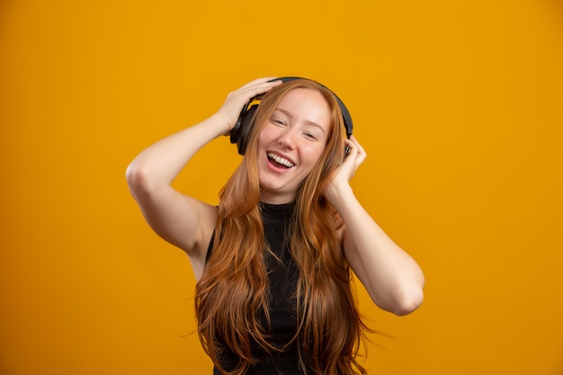 Felice e spensierata ragazza hipster rossa eccitata come le sue nuove cuffie, ballando con le mani alzate e sorridente goditi l'ascolto di musica sulla parete arancione, migliora l'umore con la canzone preferita.