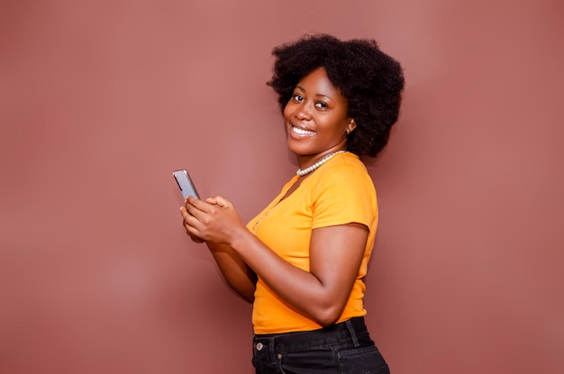 Felice eccitata signora afroamericana su uno stile di capelli afro utilizzando il telefono cellulare in studio in piedi dietro uno sfondo bianco