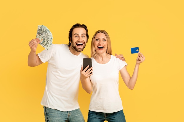 Felice eccitato uomo e donna caucasici millenari con la bocca aperta tengono la carta di credito dello smartphone e un sacco di soldi