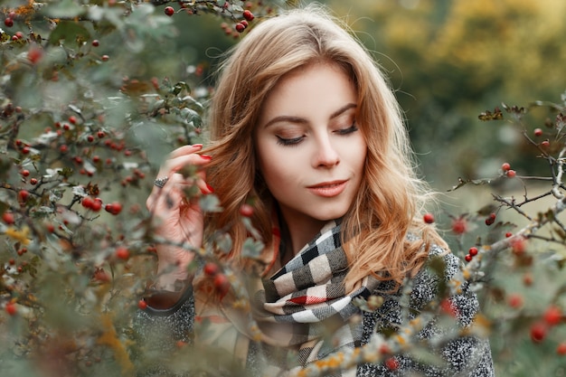 Felice elegante bella giovane donna con sciarpa a scacchi vintage in lussuoso cappotto caldo è in piedi vicino a un albero nella foresta