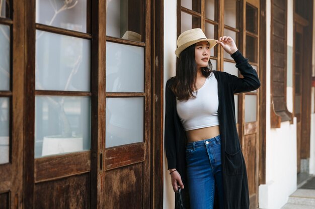 Felice elegante giovane donna coreana asiatica appoggiata al muro bianco semplice da una porta di legno a kyoto in giappone. Copia lo spazio e guarda a destra della cornice. ragazza sexy in jeans e cappello di paglia in piedi all'aperto.