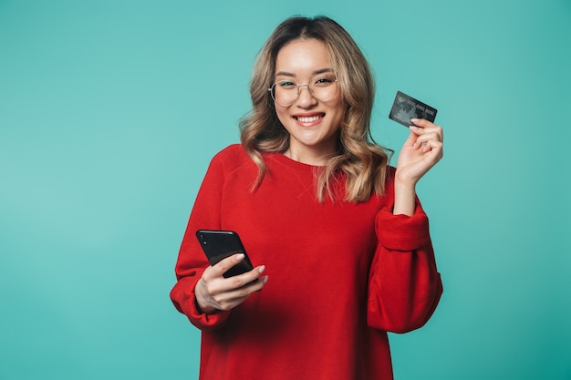 felice emotiva giovane donna in posa isolata sopra la parete blu della parete utilizzando il telefono cellulare in possesso della carta di credito