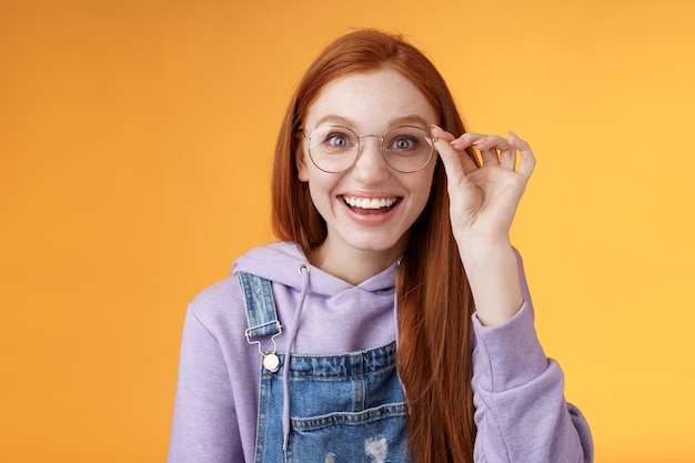 Felice entusiasta giovane ragazza rossa divertita scoprire posto eccellente celebrare b-day in piedi gioioso eccitato toccare occhiali sorridenti largamente denti bianchi sorridendo gioia sorpreso, sfondo arancione.