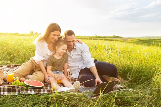 Felice famiglia amorevole che si gode un picnic insieme all'aperto, seduto su una coperta e usando il laptop, copia spazio