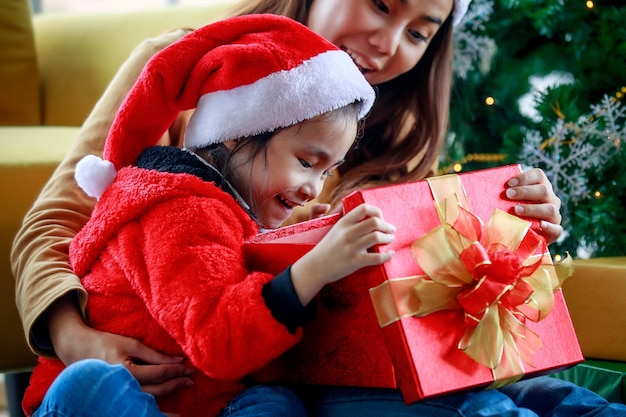 Felice famiglia asiatica figlia ragazza indossa un maglione cappello di Babbo Natale rosso e bianco seduto con la mamma unboxing regalo regalo aperto che celebra la vigilia di Natale vicino all'albero di pino di Natale nel soggiorno di casa.