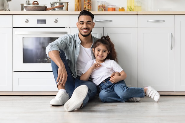 Felice famiglia mediorientale padre e figlia in posa in cucina, seduti sul pavimento e abbracciati. Allegro papà arabo e bambina che si divertono insieme a casa, panorama con spazio copia