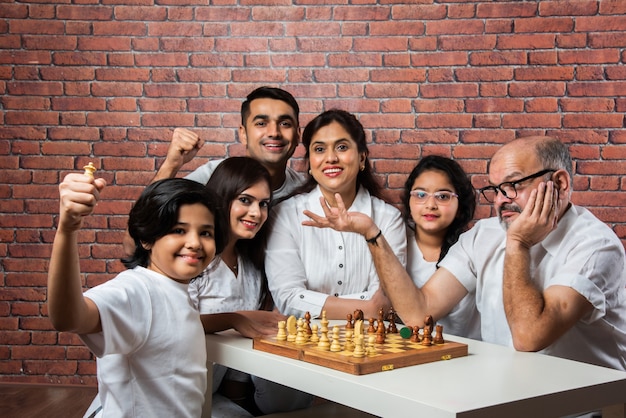 Felice famiglia multigenerazionale asiatica indiana di sei che giocano a scacchi o Shatranj che è un popolare gioco da tavolo, indossando panni bianchi contro il muro di mattoni rossi