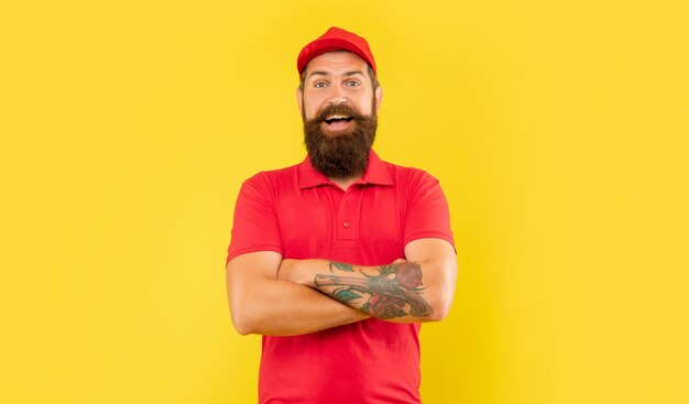 Felice fattorino fiducioso in berretto rosso casual e maglietta mantenendo le braccia tatuate incrociate sfondo giallo fiducia