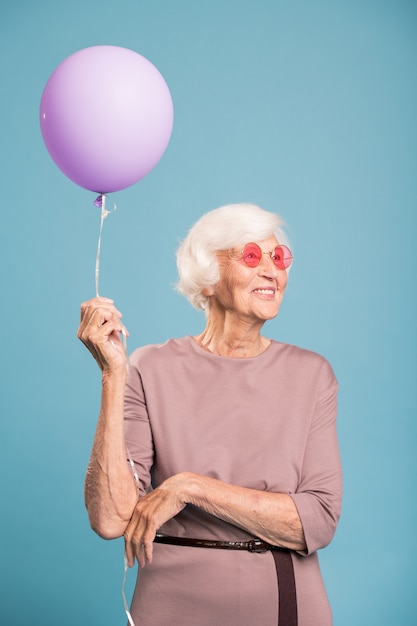 Felice femmina senior in abito elegante tenendo il palloncino mentre si sta in piedi sulla parete blu