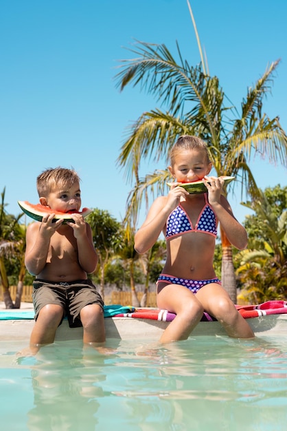 Felice fratello e sorella birazziali che mangiano anguria a bordo piscina. Trascorrere tempo di qualità, stile di vita, famiglia, infanzia, estate e concetto di vacanza.