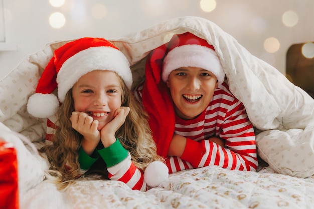 Felice fratello e sorella con i cappelli di Capodanno, giacciono sotto una coperta in pigiama di Capodanno. Natale e Capodanno.