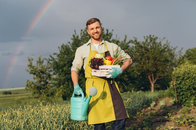 Felice giardiniere uomo che tiene cesto con verdure fresche su arcobaleno e giardino sfondo concetto di giardinaggio