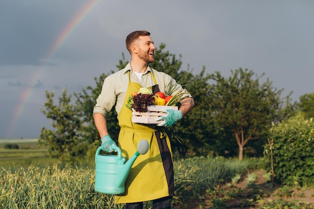 Felice giardiniere uomo che tiene cesto con verdure fresche su arcobaleno e giardino sfondo concetto di giardinaggio