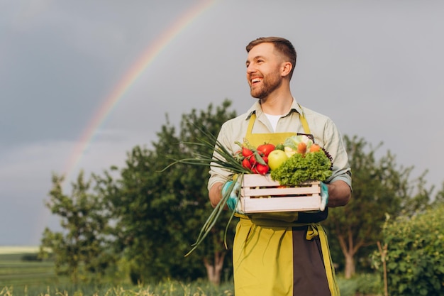 Felice giardiniere uomo che tiene cesto con verdure fresche su arcobaleno e giardino sfondo concetto di giardinaggio