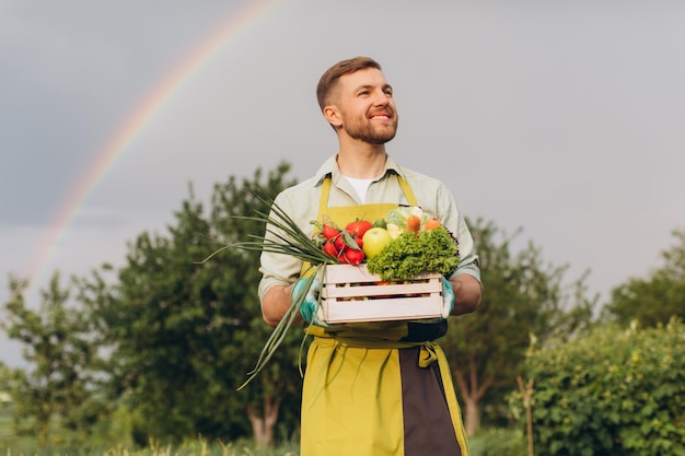 Felice giardiniere uomo che tiene cesto con verdure fresche su arcobaleno e giardino sfondo concetto di giardinaggio