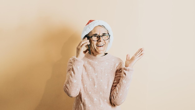 Felice gioiosa anziana vecchia capelli bianchi donna che indossa il cappello di Babbo Natale sorridente durante l'utilizzo di smart phone. si erge su uno sfondo colorato. Indossare abiti natalizi. Copia spazio. Spazio pubblicitario