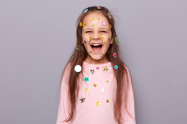 Felice gioiosa bambina allegra coperto adesivi colorati per bambini in piedi isolato su sfondo grigio e ridendo essendo di buon umore giocoso esprimendo emozioni positive