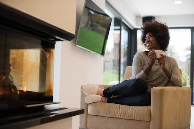 Felice gioiosa donna afroamericana che beve una tazza di caffè rilassante al camino. Giovane ragazza nera con riscaldamento di bevande calde in fase di riscaldamento. autunno a casa.